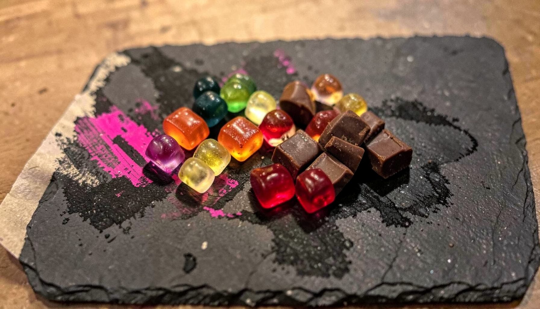 Assorted colourful gummies and chocolate squares arranged on a dark slate board, warm ambient light